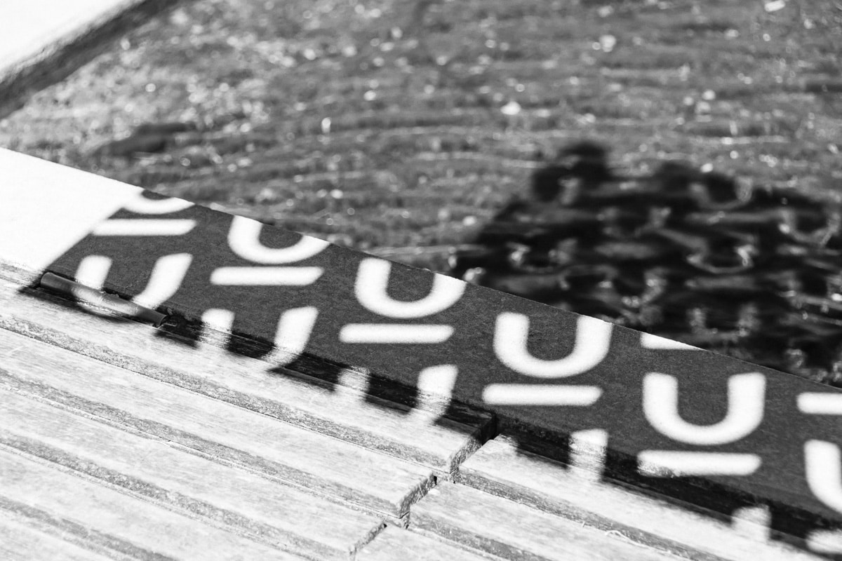 Close-up black and white abstract photo of layered cardboard with bold white lettering on dark tape, chain in background, Nu